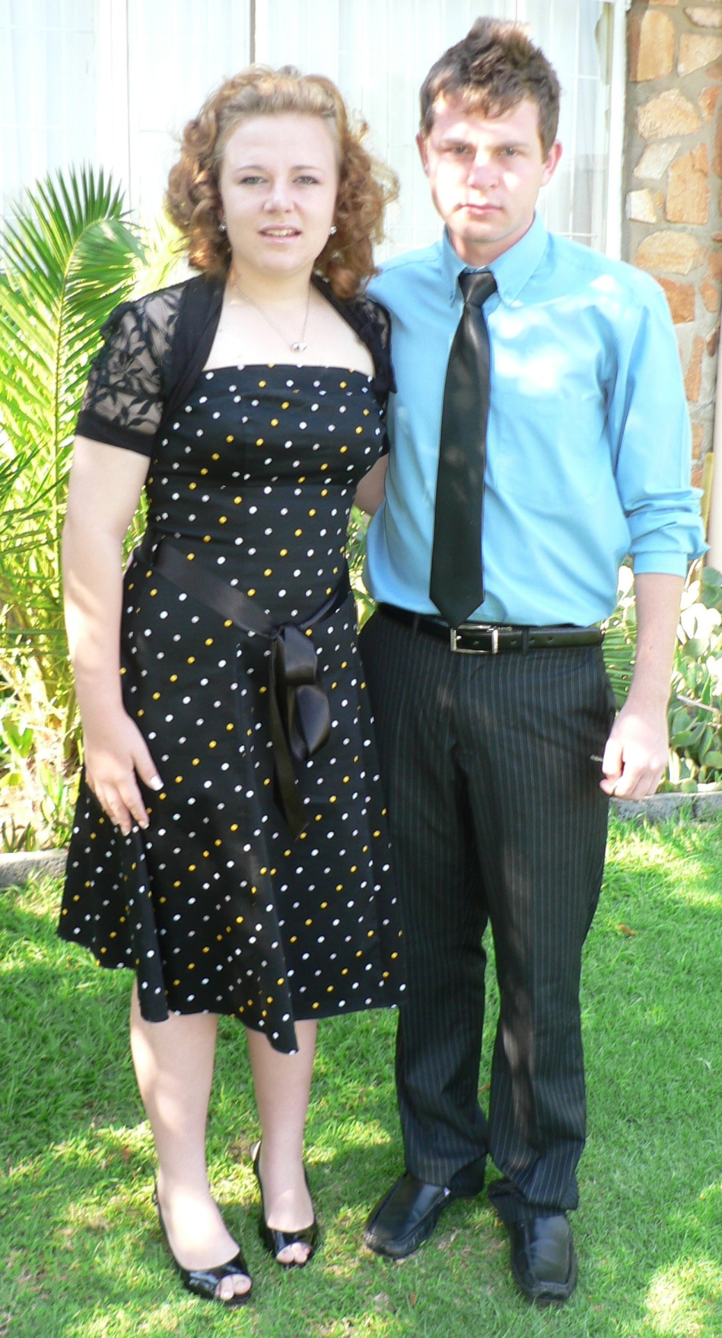 Angelo in a blue shirt and tie standing with a young woman in a polka-dot dress on the lawn