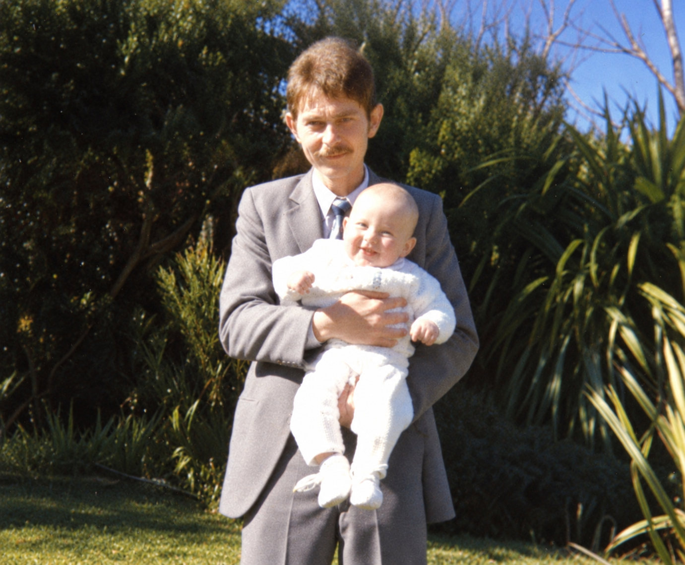 A man in a suit holding a baby in a white christening outfit among garden bushes
