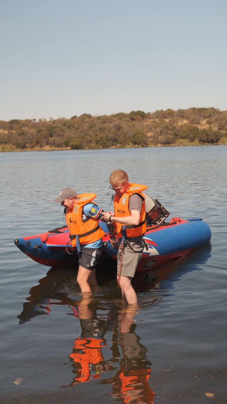 Two men in orange life vests beside an inflatable boat on the lake shore