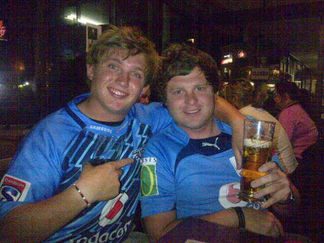 Two young friends in blue Bulls rugby jerseys sharing a beer at a pub at night