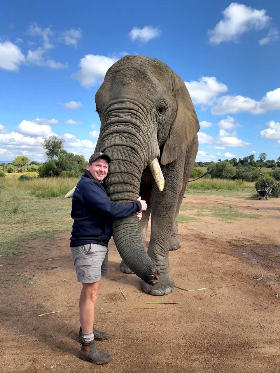 Angelo embracing the trunk of a large African elephant on an open plain