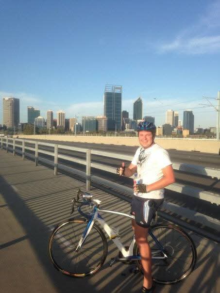 Angelo on his bicycle in helmet and cycling kit, giving a thumbs-up with a city skyline behind him