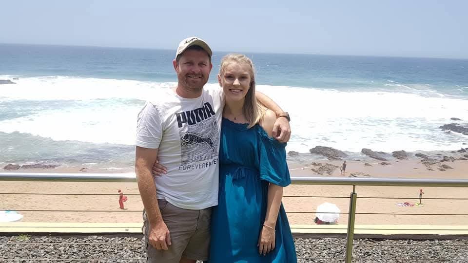 Angelo in a Puma shirt standing arm in arm with a friend in a blue dress above a beach