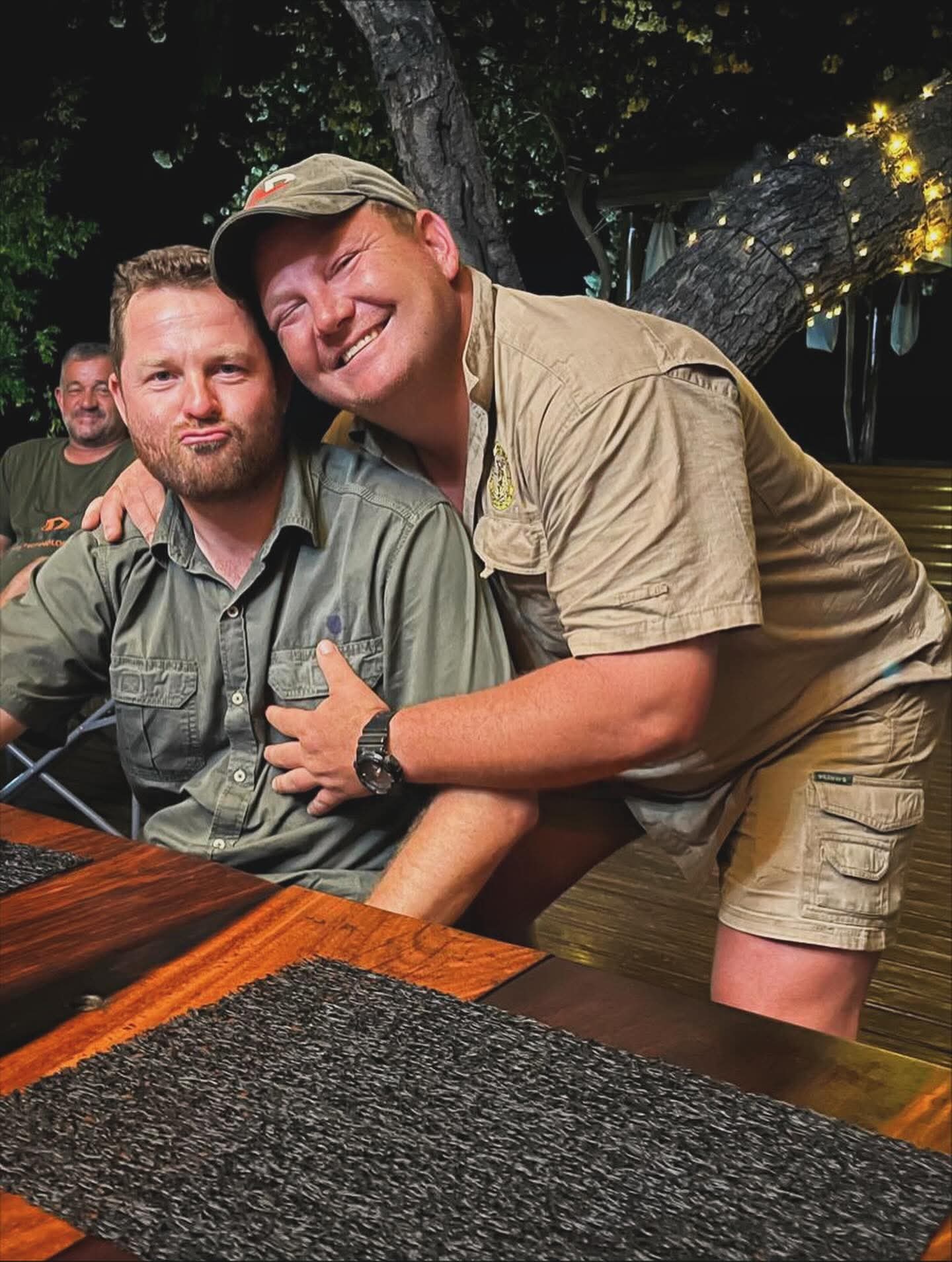Angelo hugging a smiling friend in khaki shirts at a camp table at night