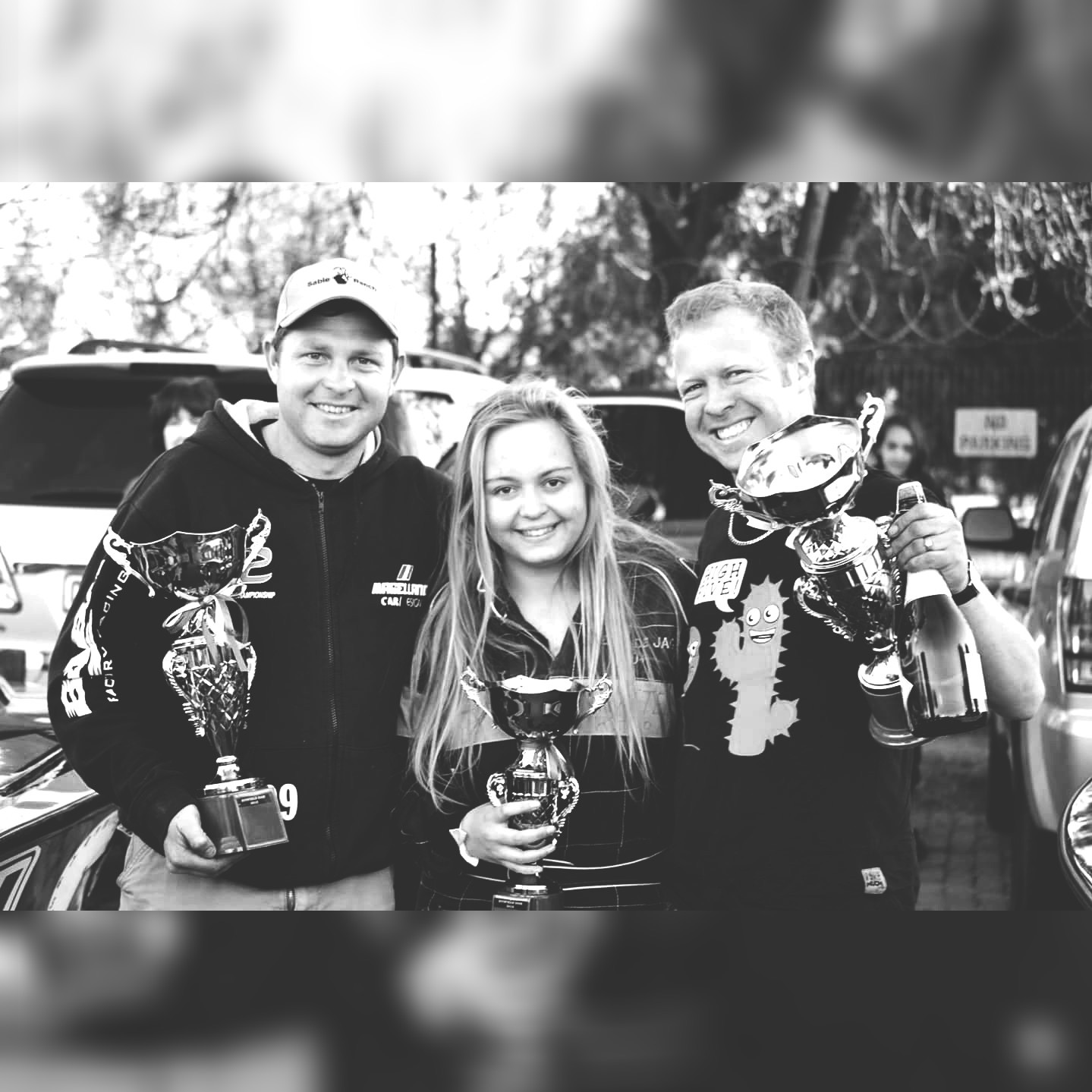 Three smiling friends holding racing trophies after a rally, in black and white