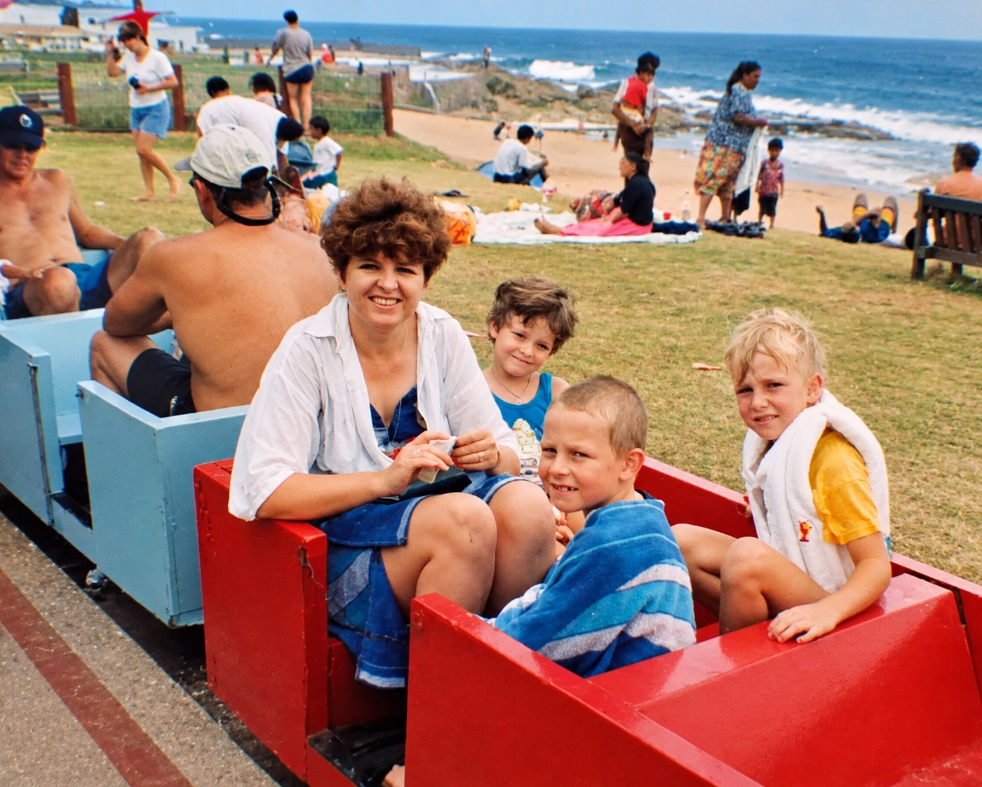 A woman and three boys riding a small red train along a coastal path beside the beach