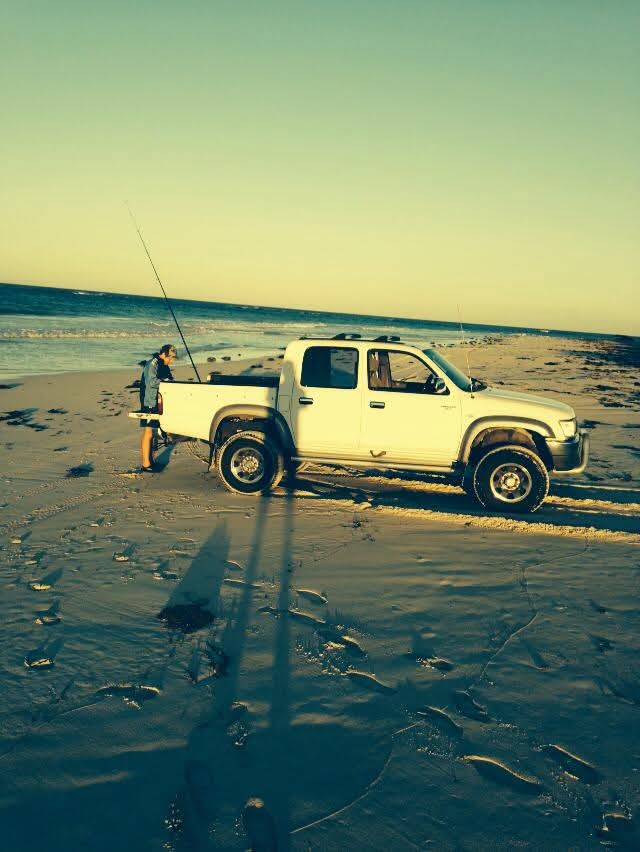A white bakkie parked beside the ocean with fishing rods and a person standing at the tailgate in golden light