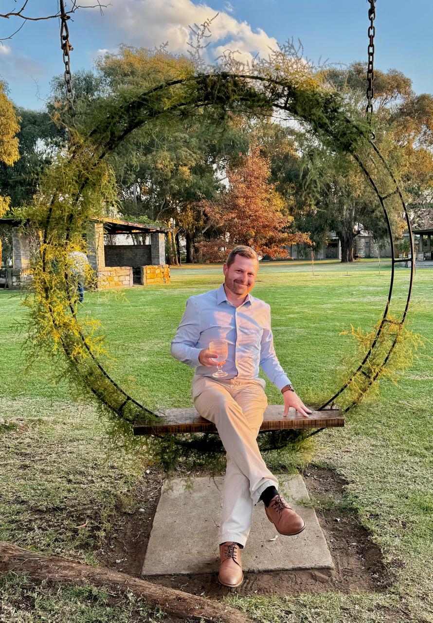 Angelo sitting relaxed on a garden swing seat holding a glass, smiling at golden hour