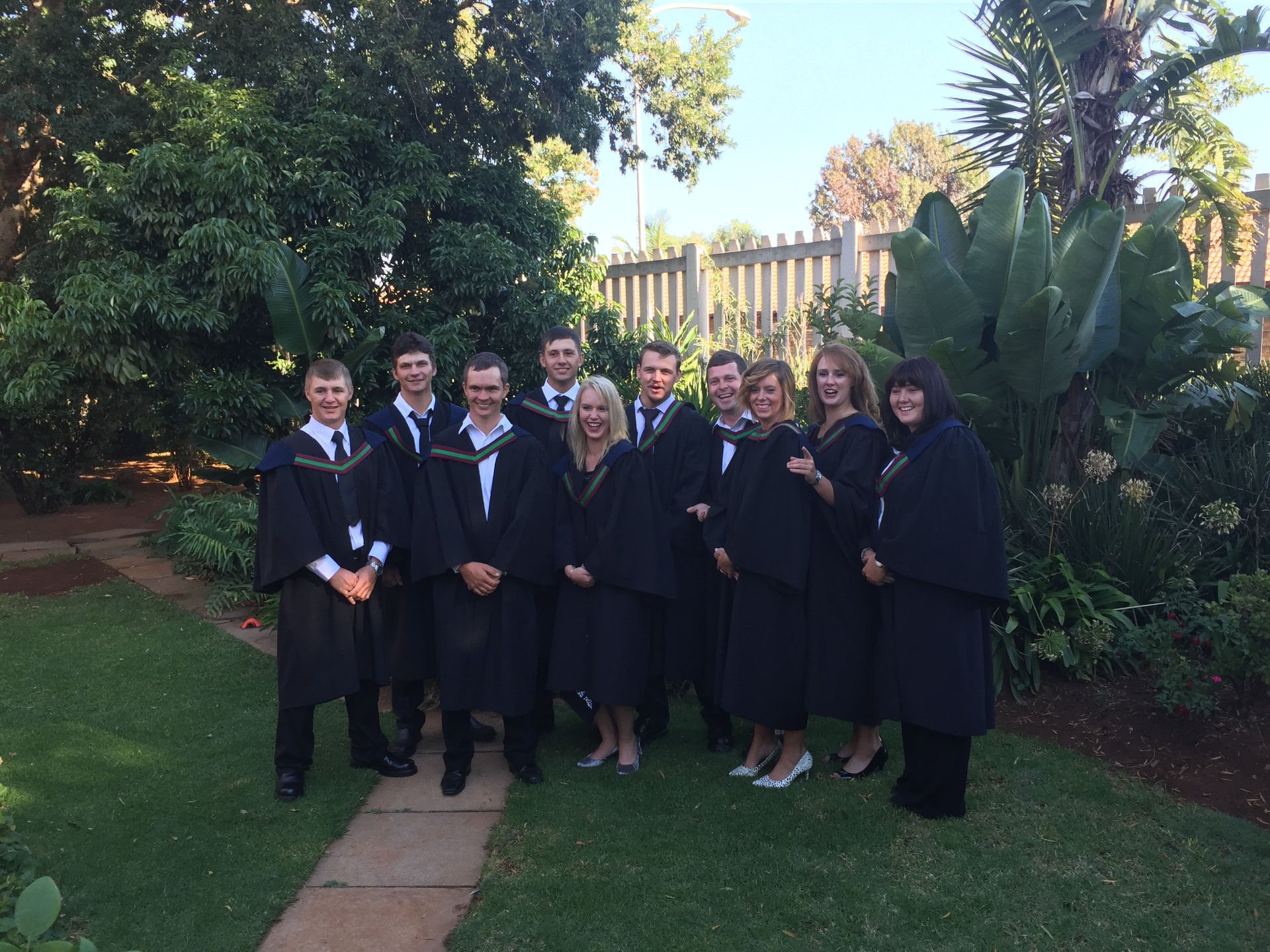 A group of graduates in black gowns with green and red sashes laughing together in a garden