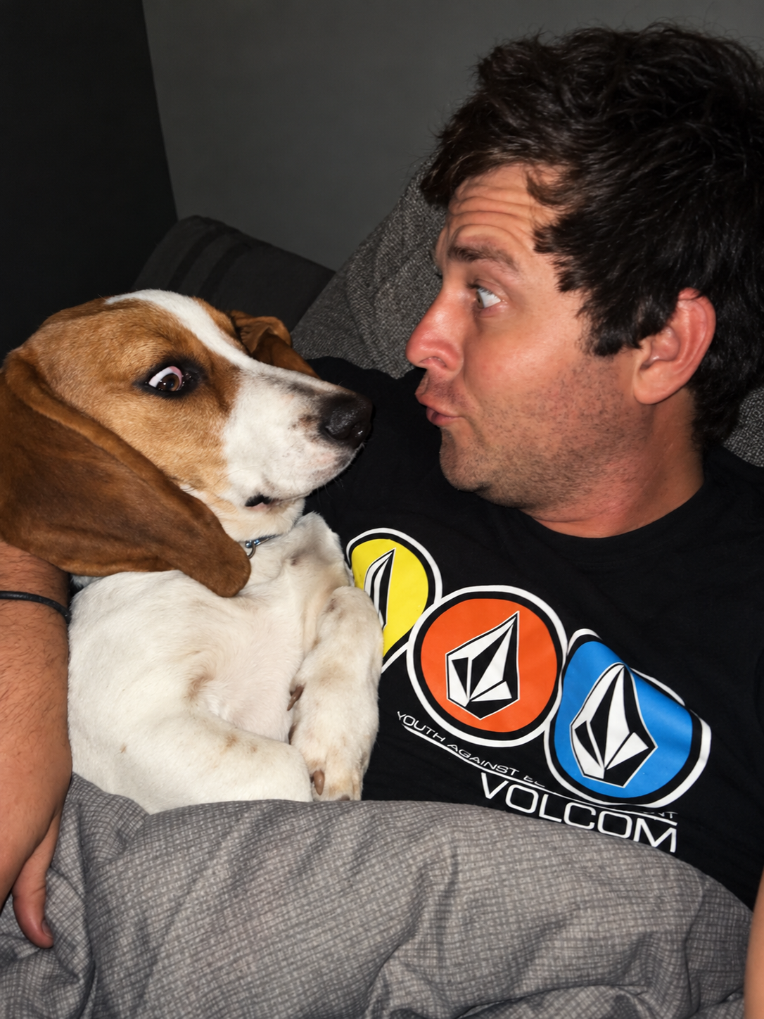 Angelo tucked under a blanket on the couch beside his basset hound, both looking at each other with wide-eyed, playful faces