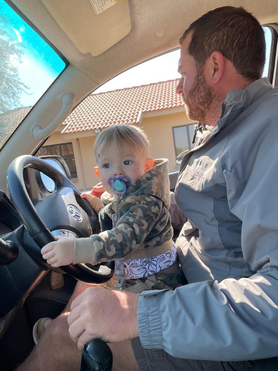 A toddler with a dummy holding the steering wheel on a man's lap