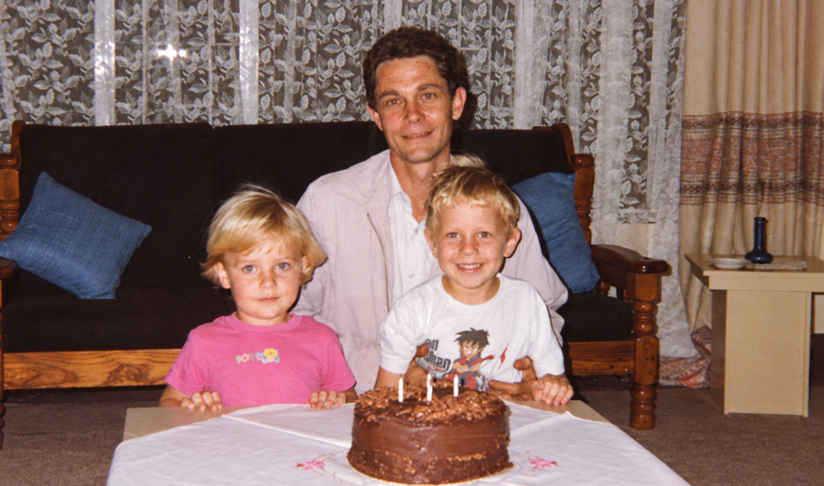 A man and two children seated behind a chocolate birthday cake with lit candles
