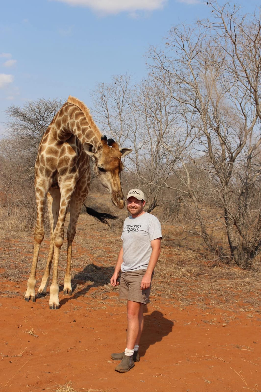 Angelo standing and smiling in a dry bushveld clearing with a tall giraffe's head hanging right beside his