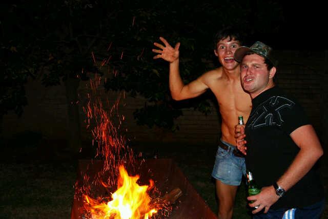 Two friends laughing beside a crackling braai fire sending sparks into the night