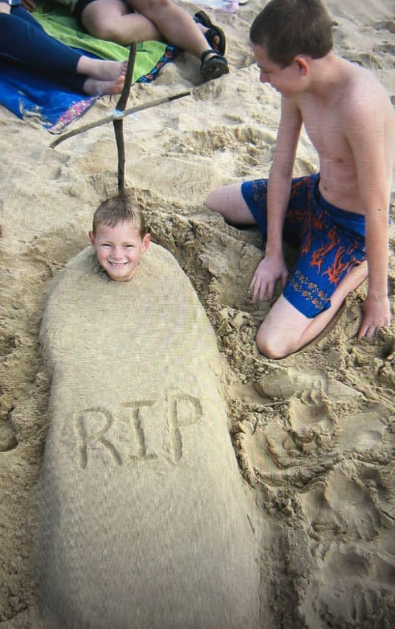 Two young boys playing on the beach, one smiling while buried in sand and the other kneeling beside him