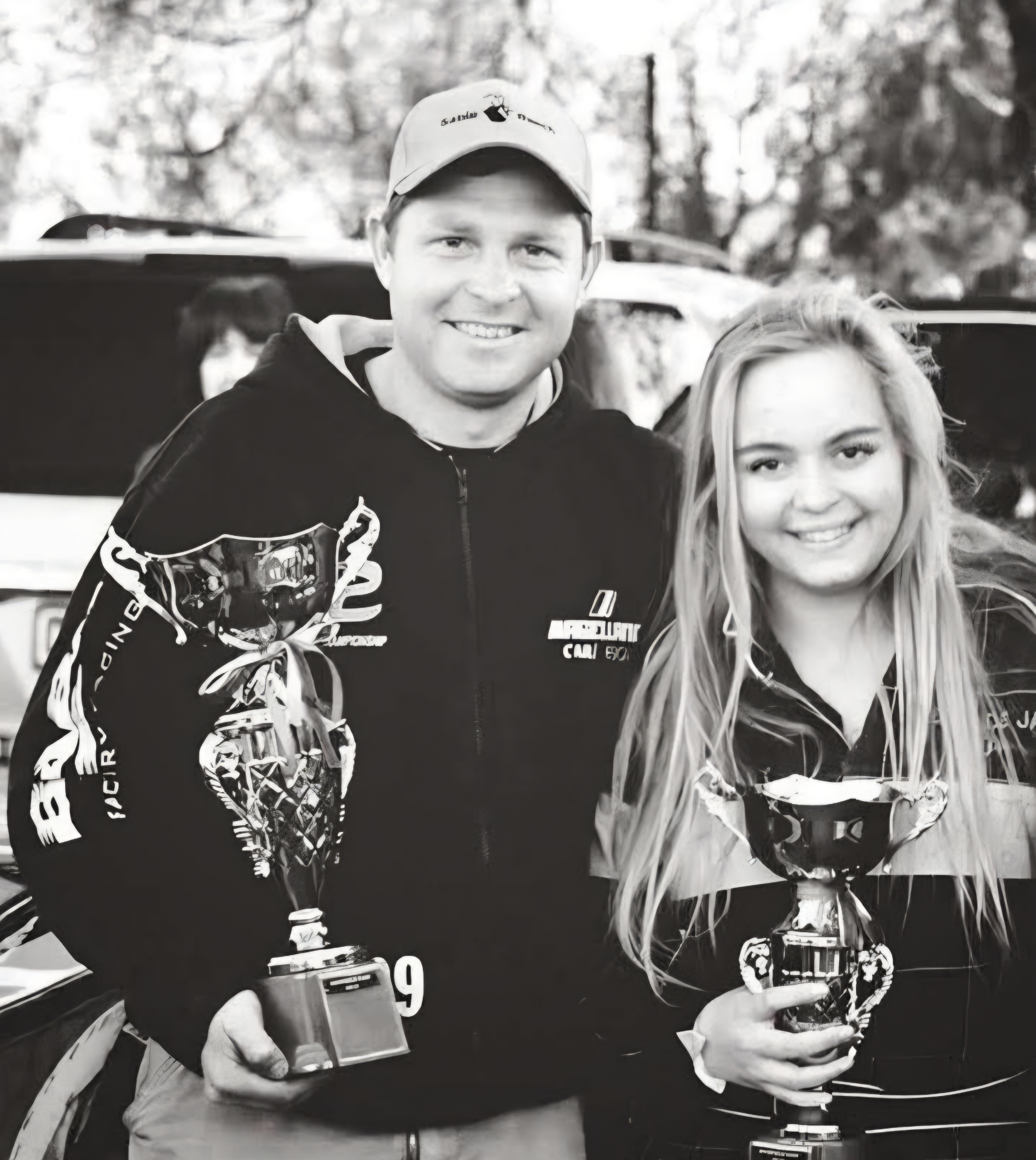 Black and white portrait of Angelo and a young woman each holding a trophy beside a car