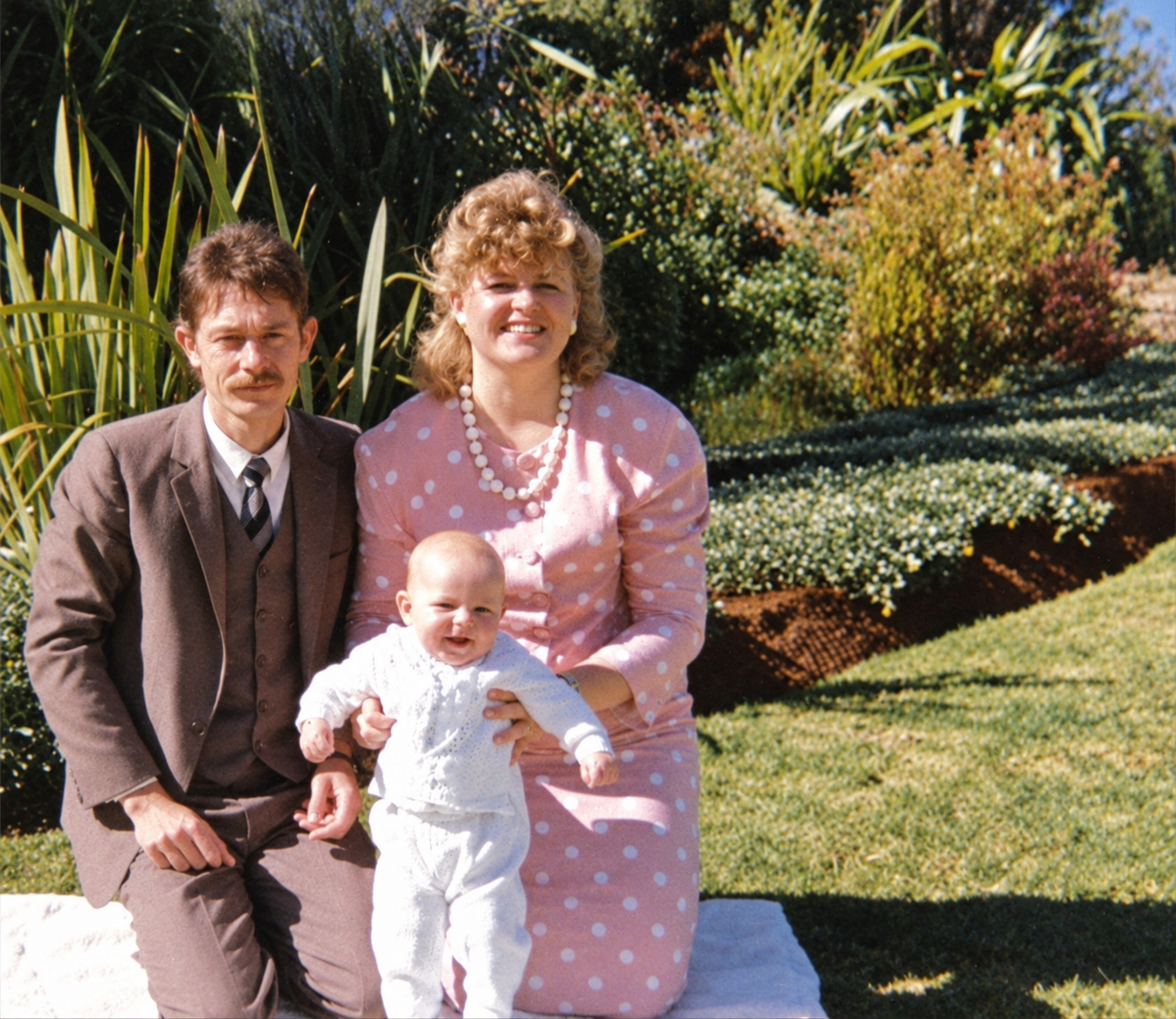 A couple kneeling on a white blanket on the lawn with a baby in a white christening outfit