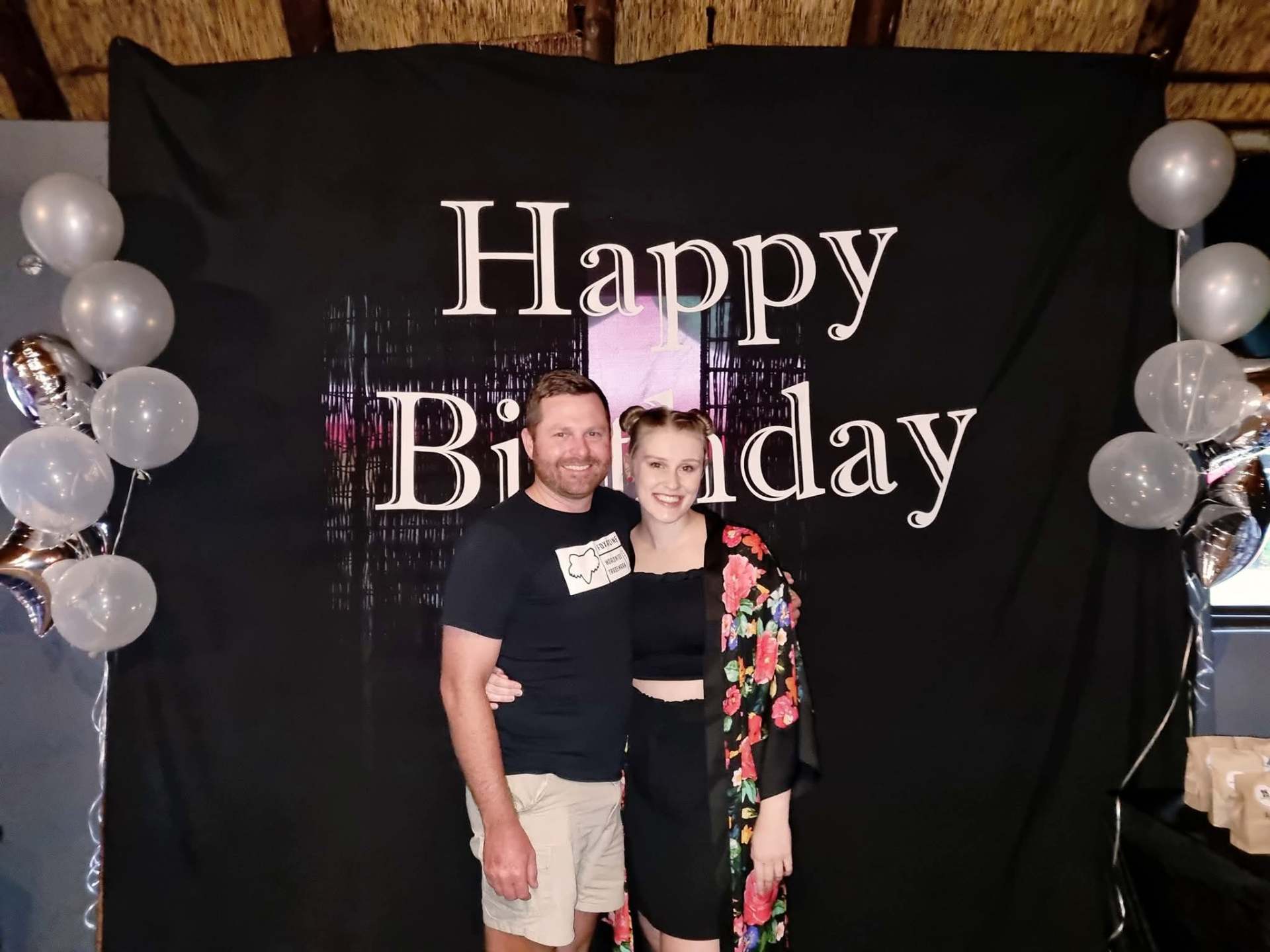 Angelo and a friend posing in front of a Happy Birthday backdrop with balloons
