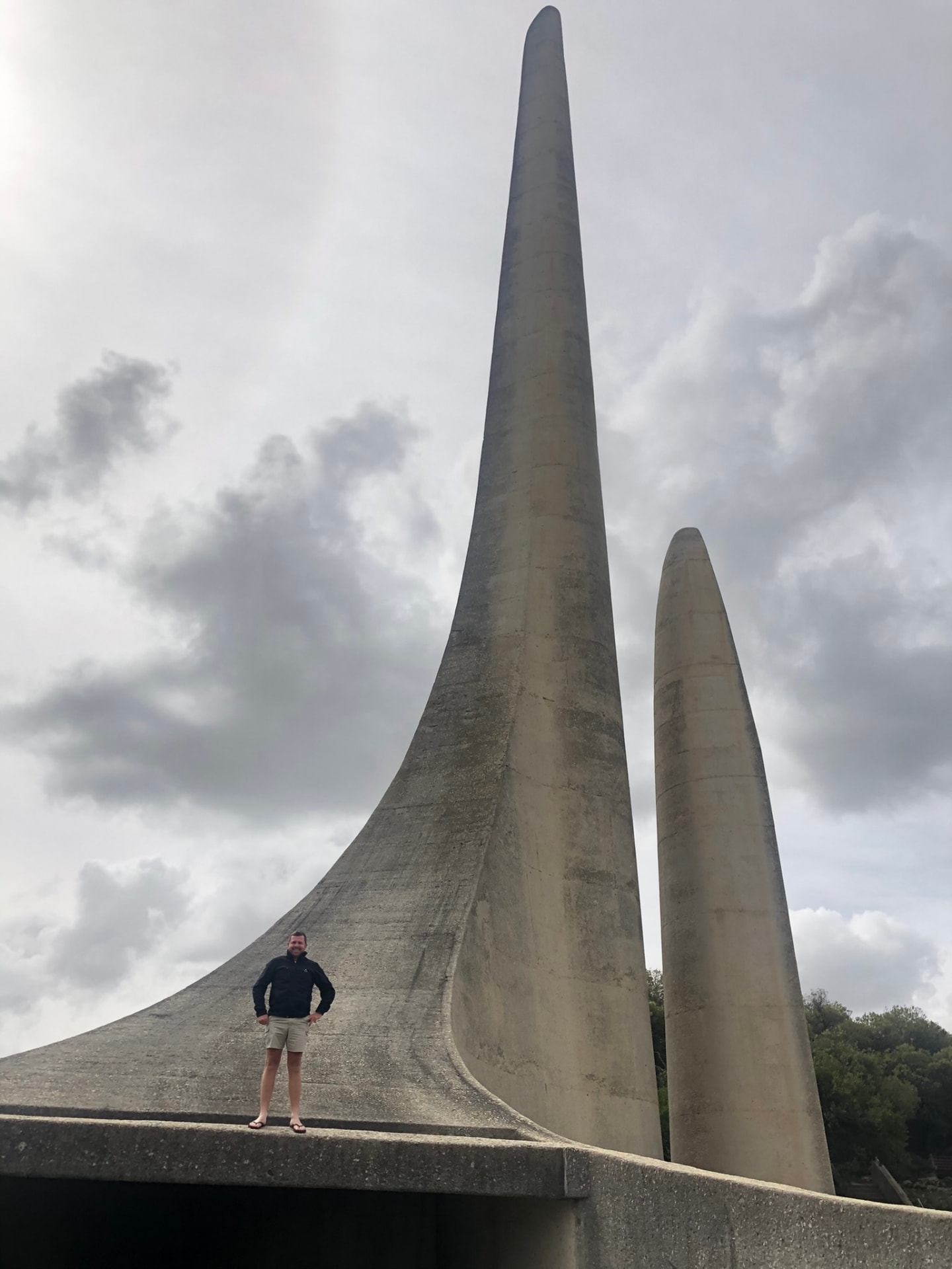 Angelo standing beside the tall curved towers of a monument beneath a cloudy sky