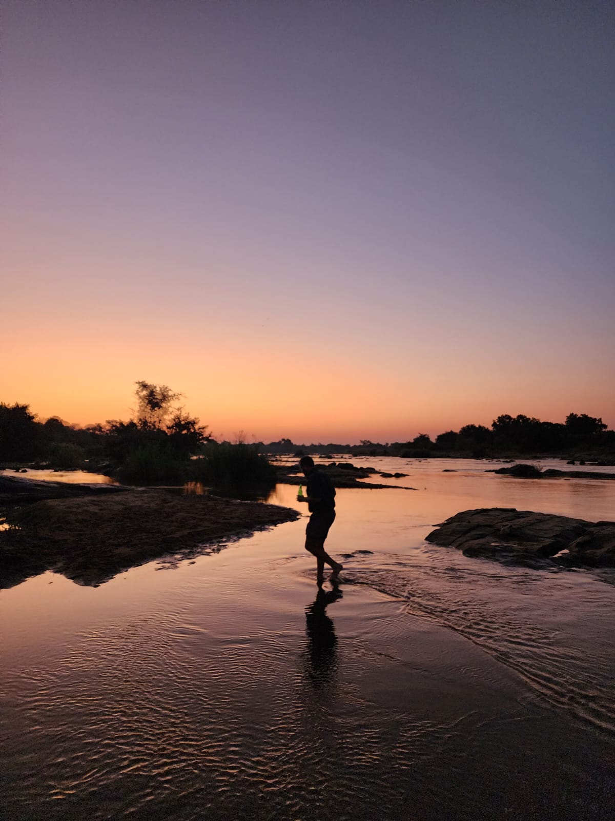 Angelo wading barefoot through a glowing river at dusk, his reflection on the water