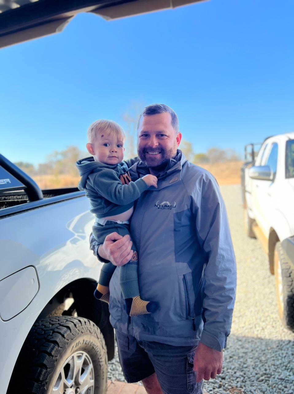 A man holding a smiling toddler beside a white bakkie under a blue sky