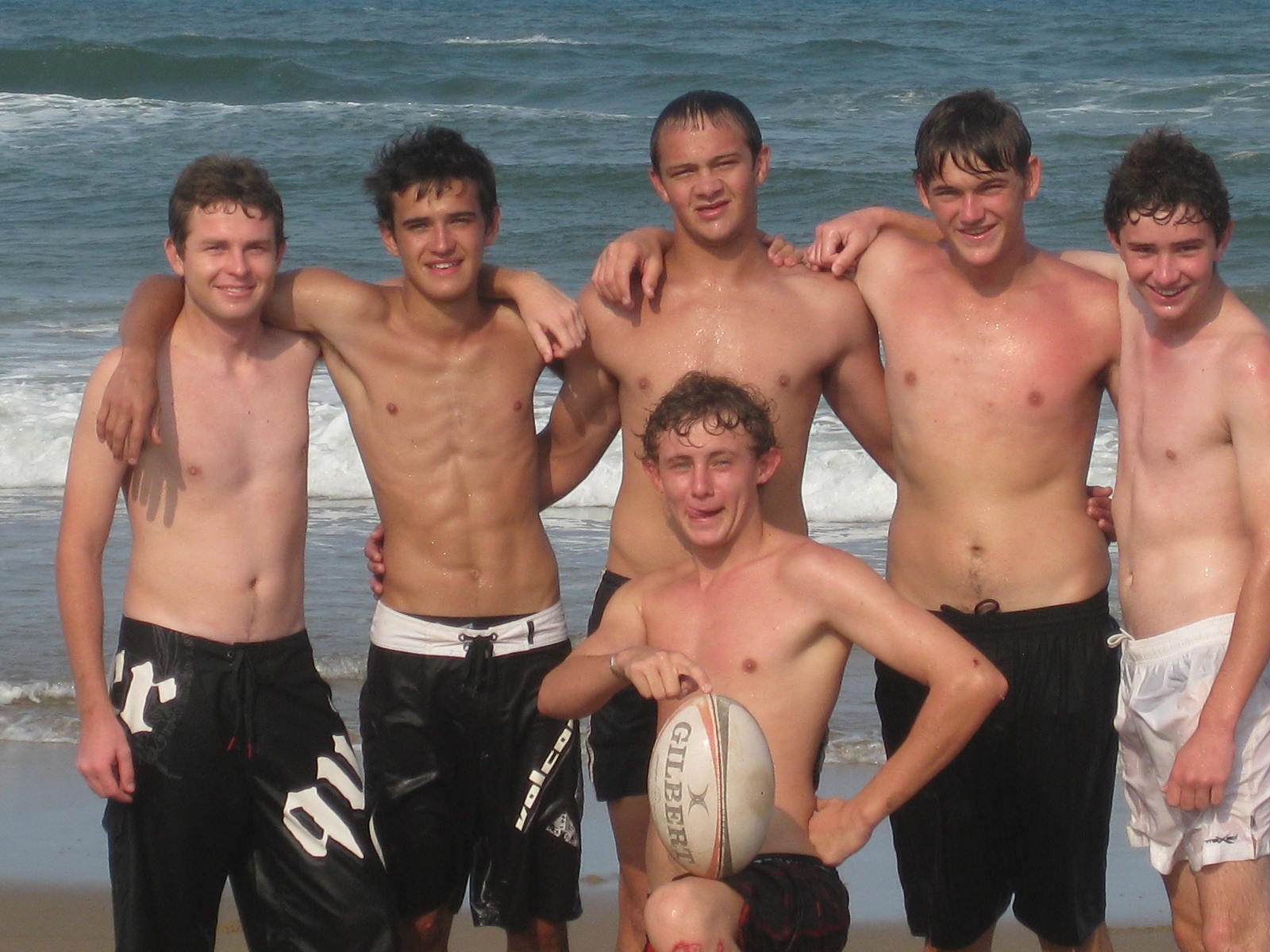 Six young friends in board shorts on the beach with a rugby ball, fresh from the waves