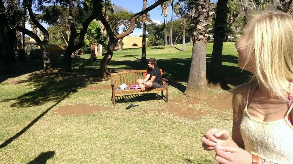 Angelo sitting on a wooden bench under palm trees in a sunlit park