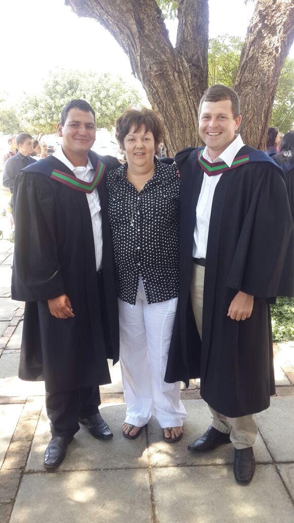 Two graduates in gowns standing with their mother under a tree on graduation day