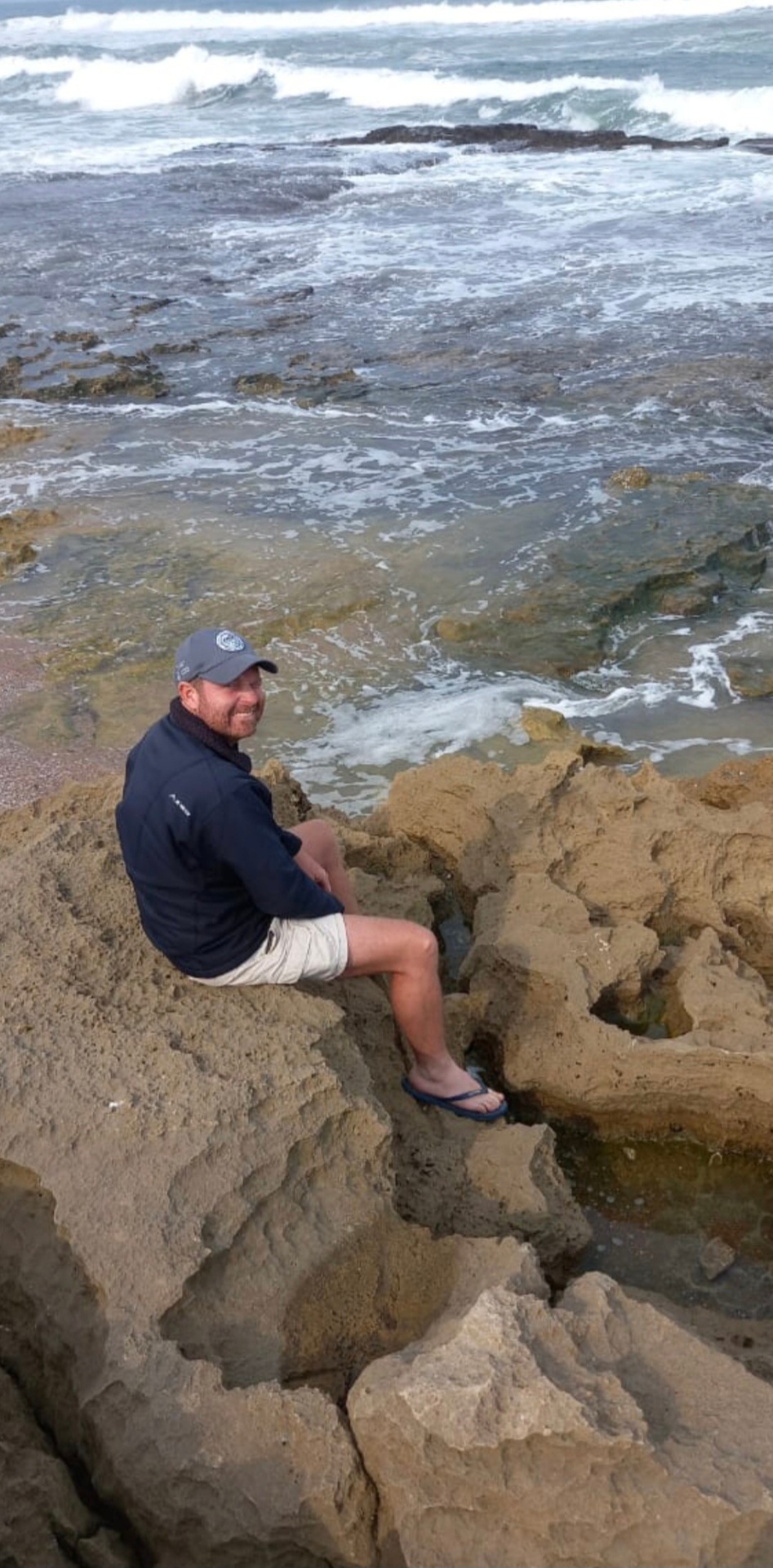 Angelo sitting on rocks overlooking a choppy ocean, smiling back at the camera