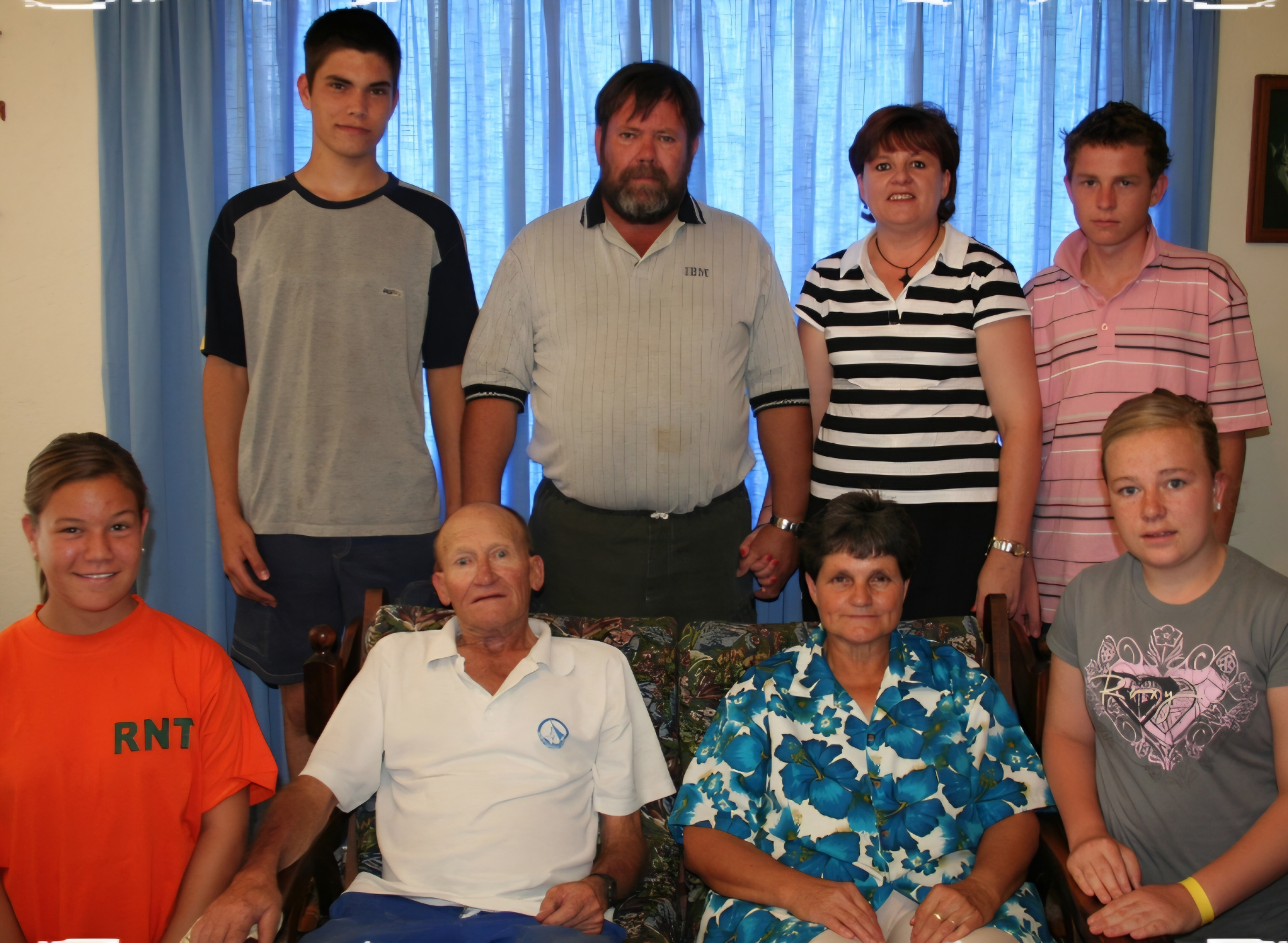 An extended family portrait with four relatives standing behind grandparents and two children seated beside them, against blue curtains