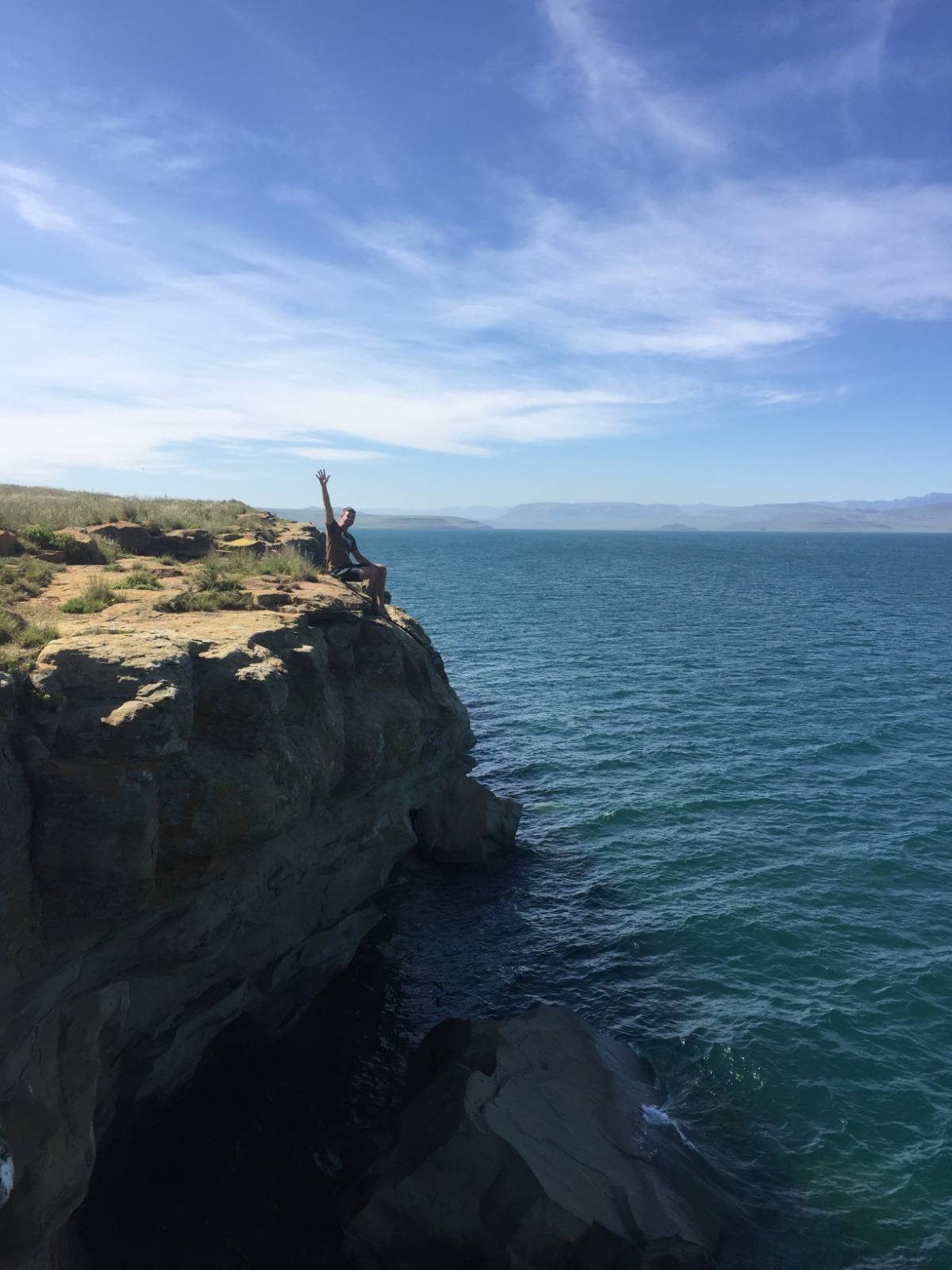 Angelo seated on a rocky cliff edge above turquoise water, waving to the camera