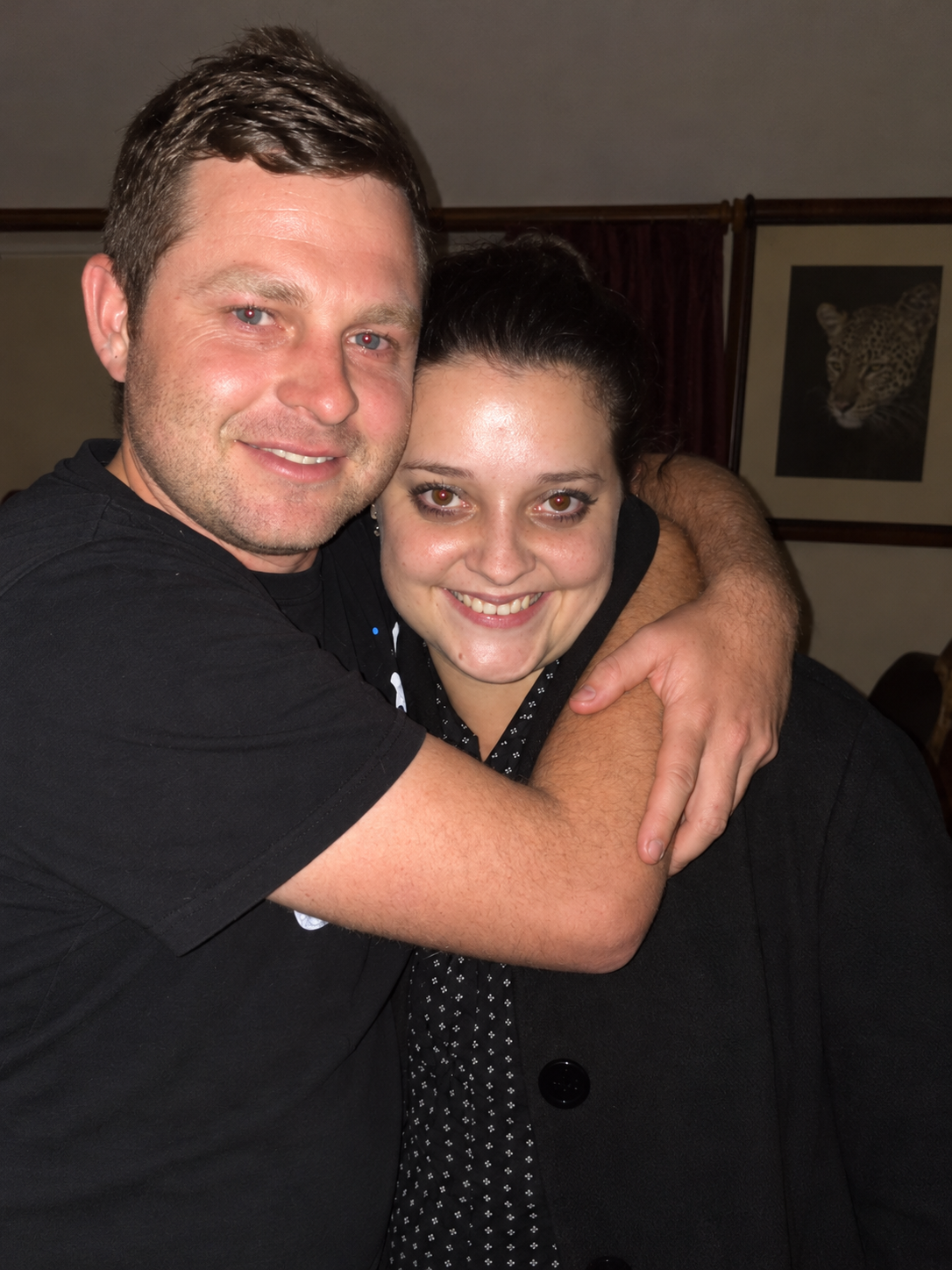 Two people in black smiling as they embrace indoors, a framed leopard print hanging on the wall behind them