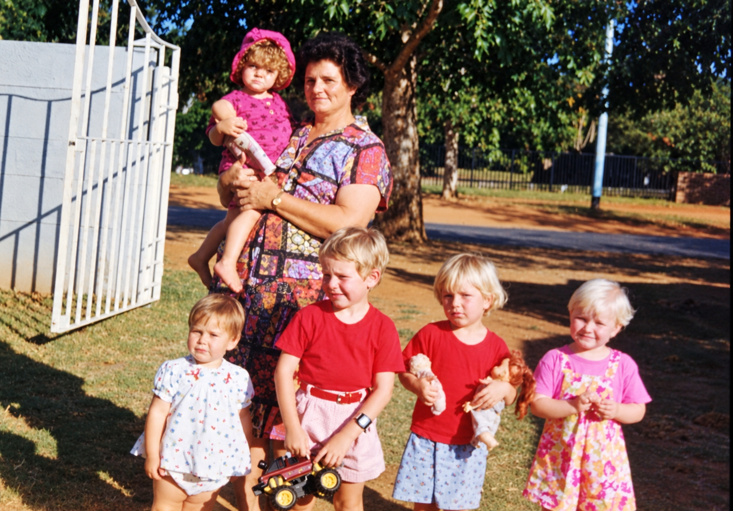 A woman holding a toddler in a pink bonnet with four young children standing in a row outdoors in front of an open white gate