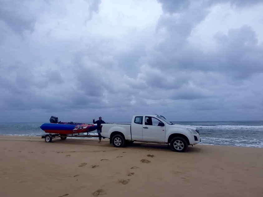 A white bakkie towing a red-and-blue inflatable boat on the beach beneath cloudy skies
