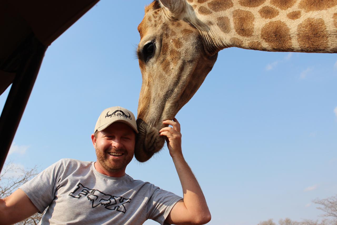 Angelo smiling in an open game-viewer vehicle as a giraffe rests its muzzle gently against his head