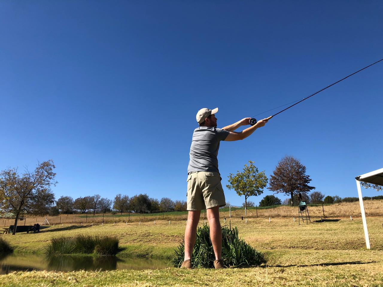 Angelo casting a fly-fishing line beside a farm dam under a deep blue sky