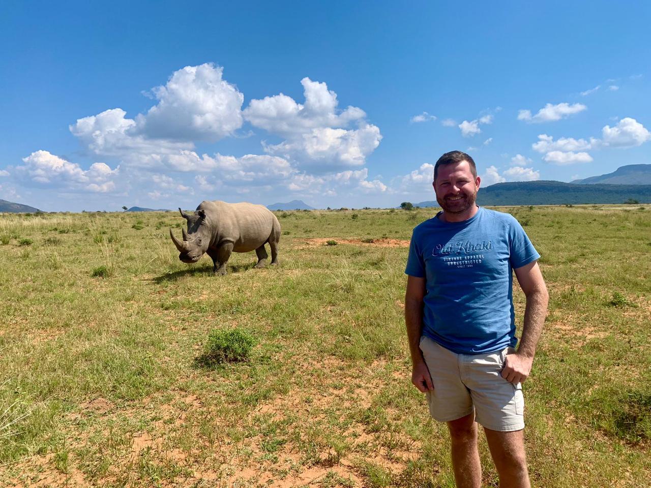 Angelo standing in open grassland with a rhinoceros nearby