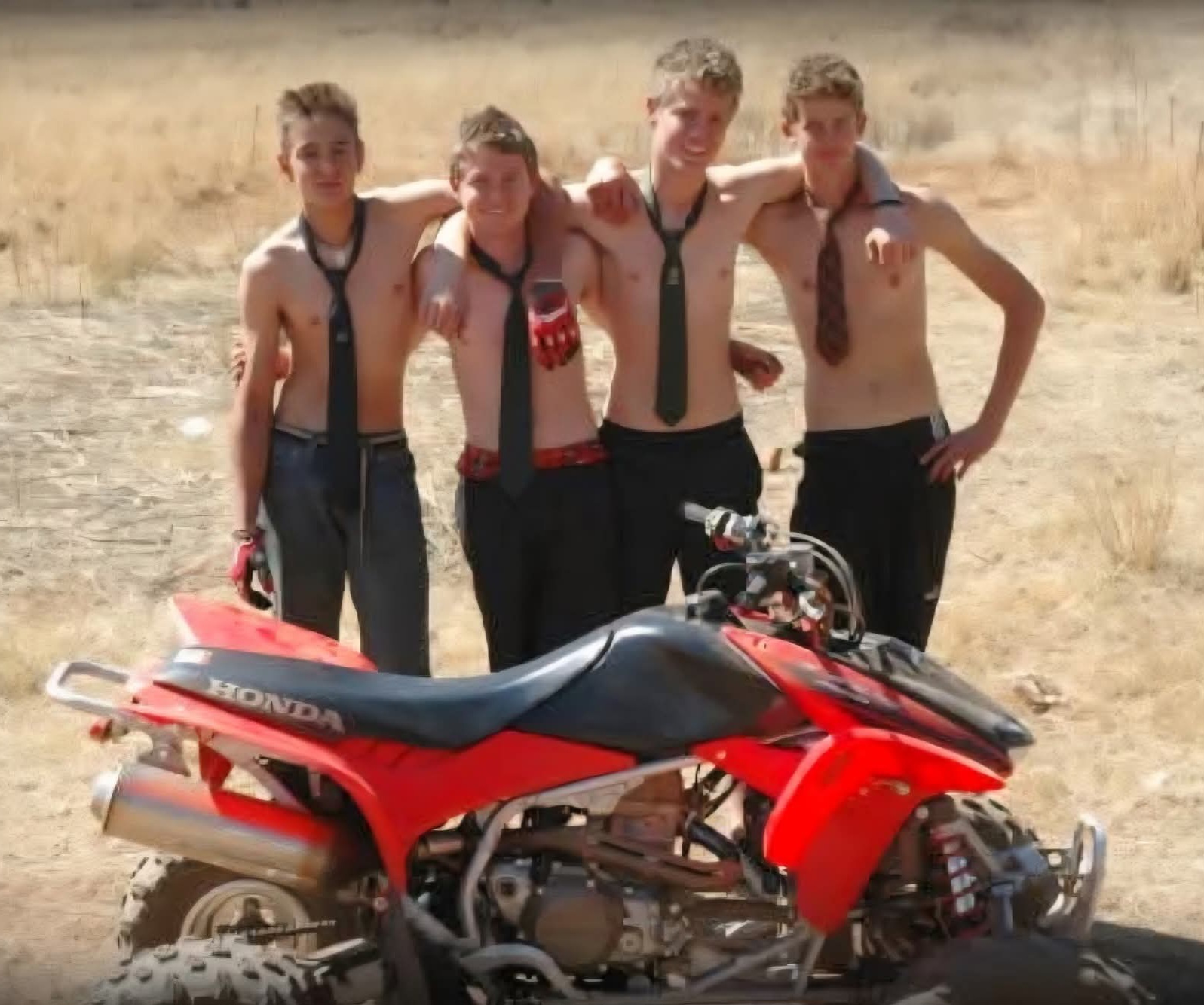Four shirtless young men wearing school ties posing beside a red Honda quad bike in dry veld