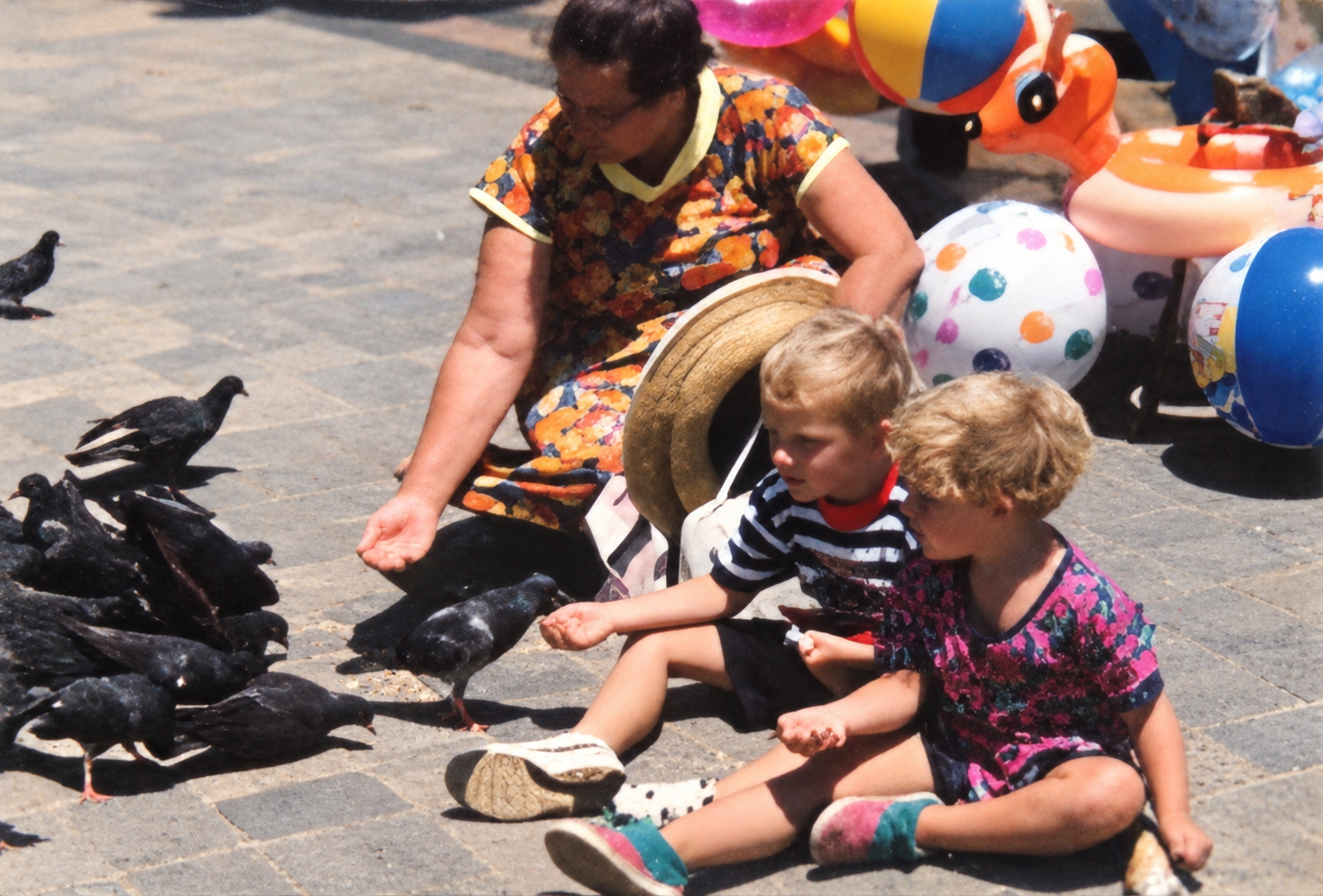A woman and two young boys sitting on a stone pavement feeding a flock of pigeons