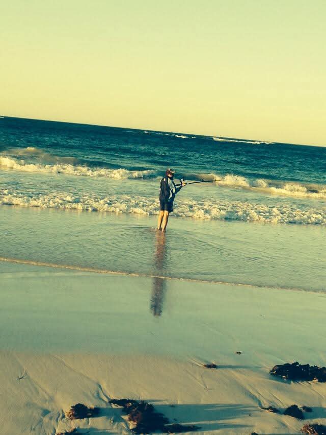 A person fishing in the shallow surf with waves rolling in and long shadows across the sand