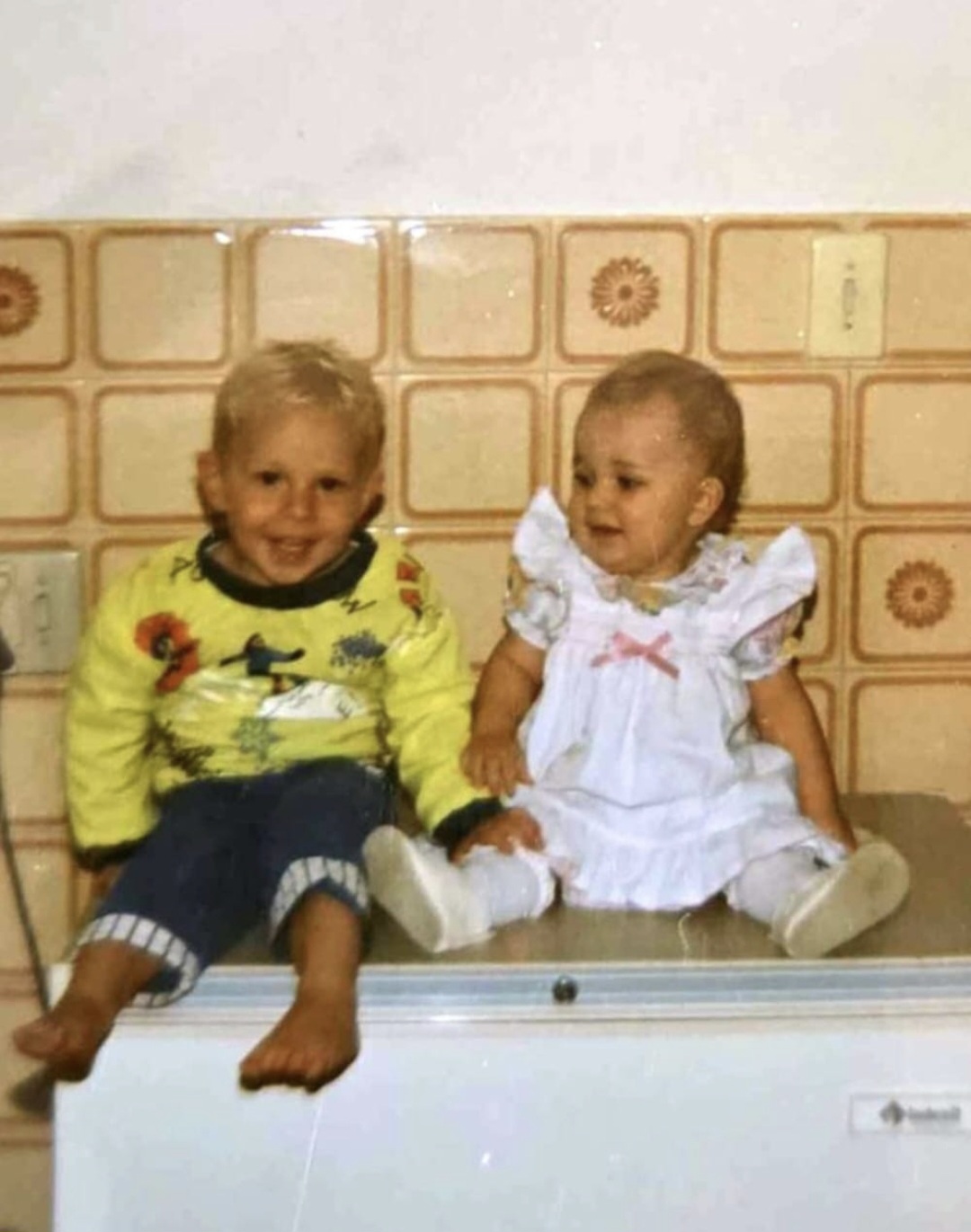 Angelo and his baby sister sitting together in a tiled kitchen