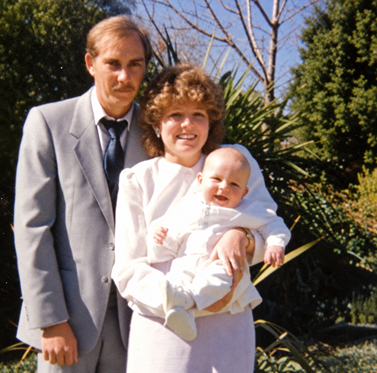 A family in formal clothes posing outdoors with a baby in a white christening outfit