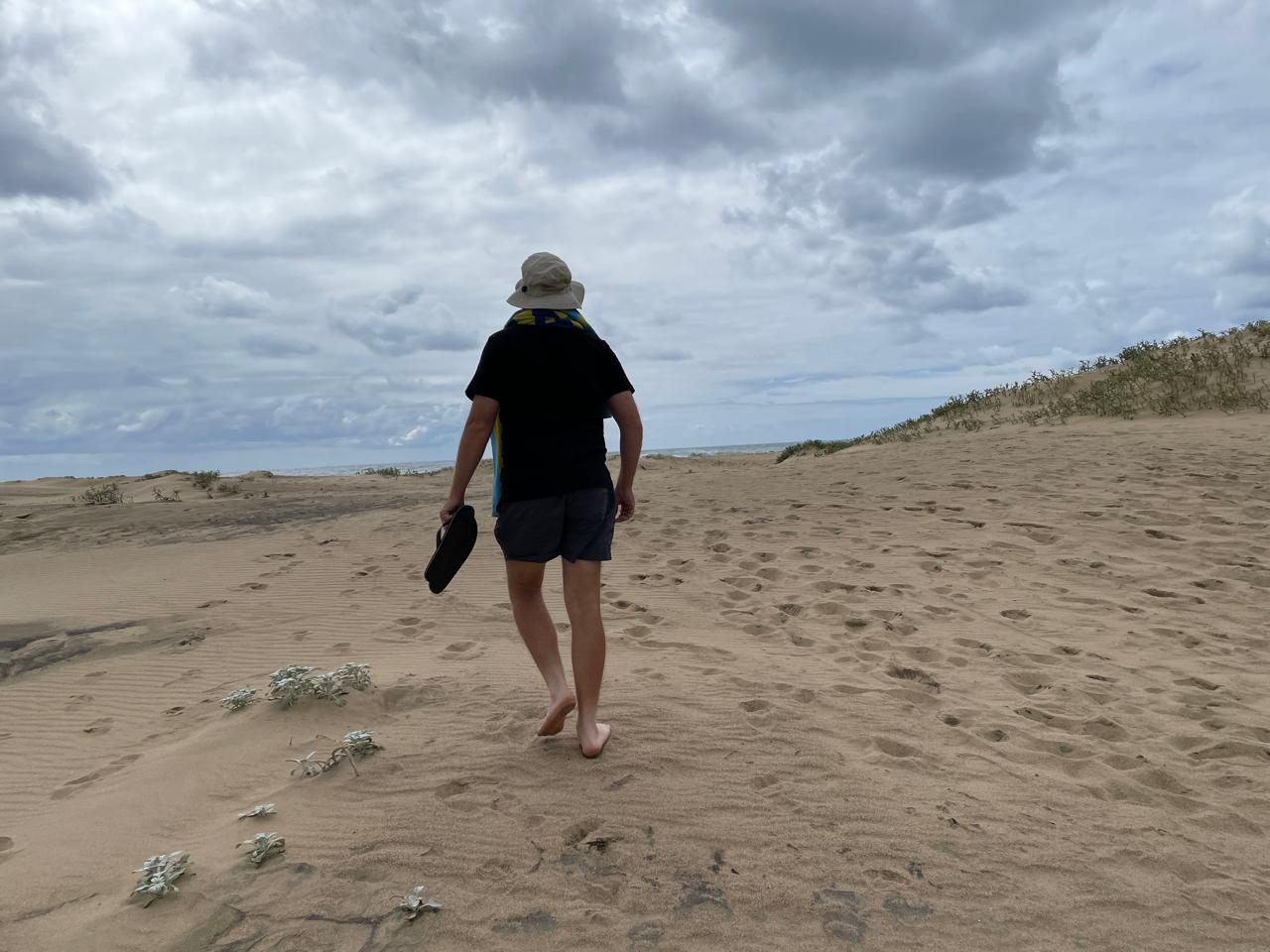 Angelo walking barefoot across the coastal dunes with his sandals in hand