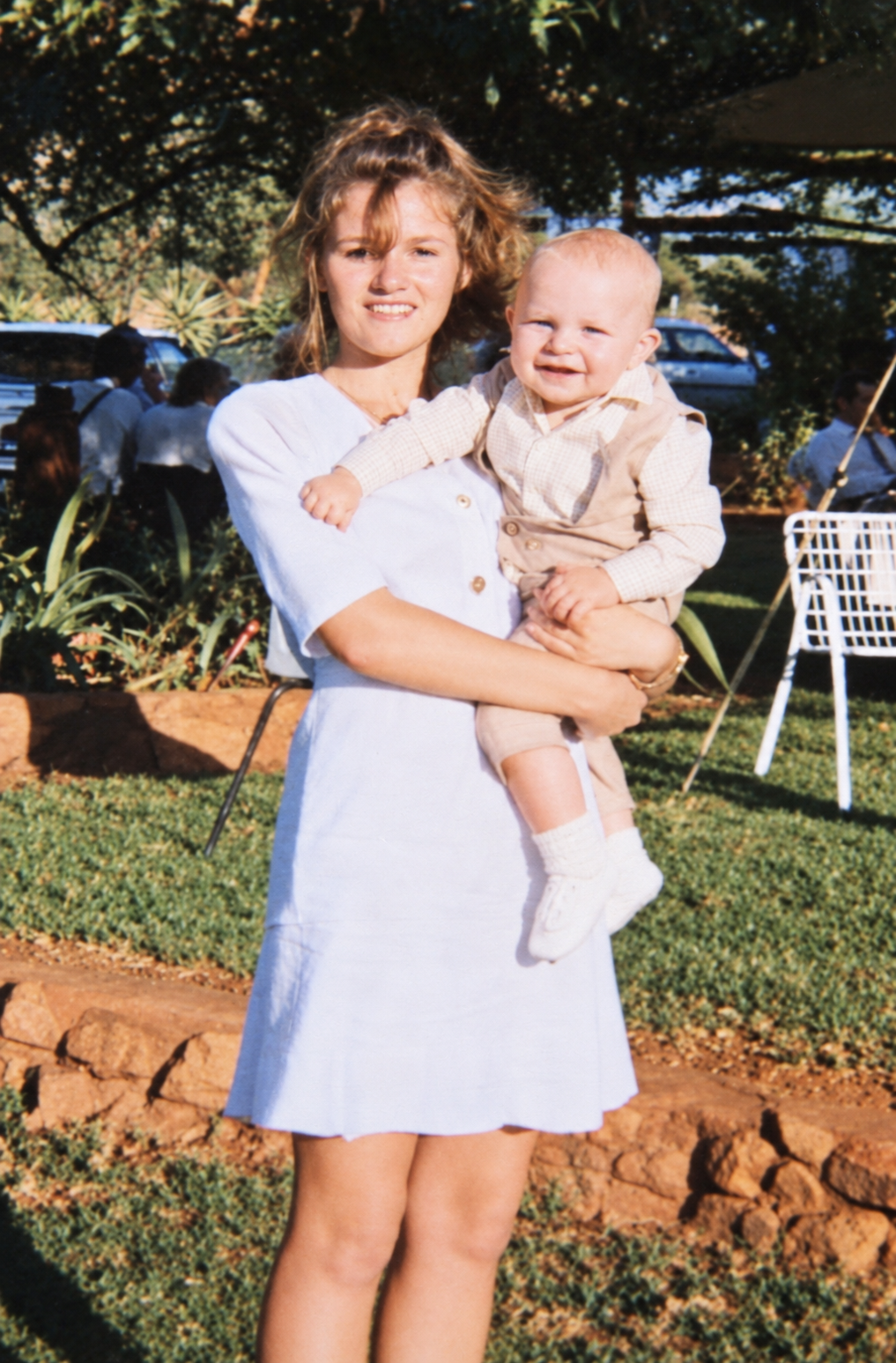 A young woman holding a smiling baby in a little waistcoat on a sunny lawn