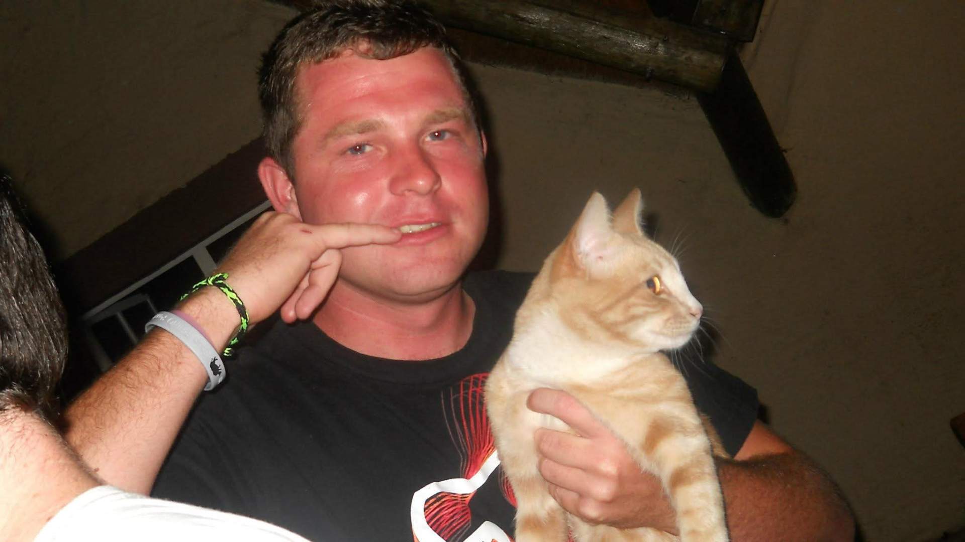 Angelo holding a ginger cat at night while a friend points playfully beside him