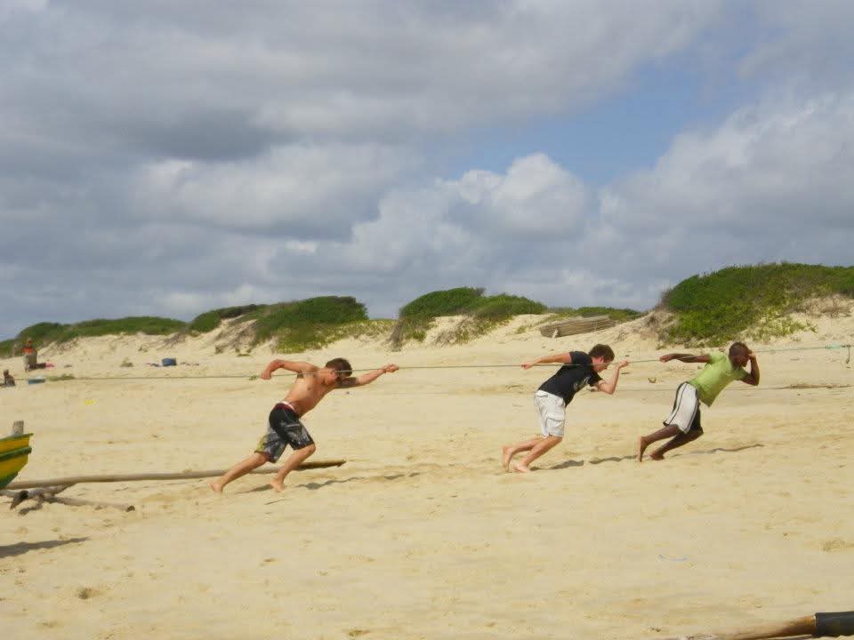 Three friends hauling a net rope along the beach with dunes behind