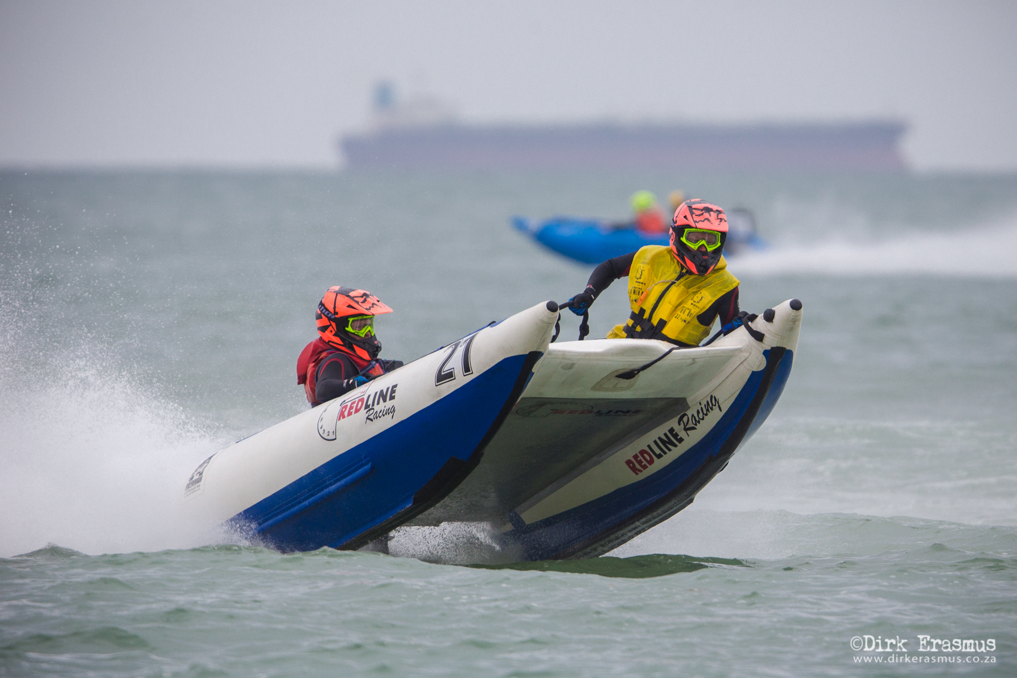 Two helmeted racers lifting a white-and-blue Redline boat over ocean waves