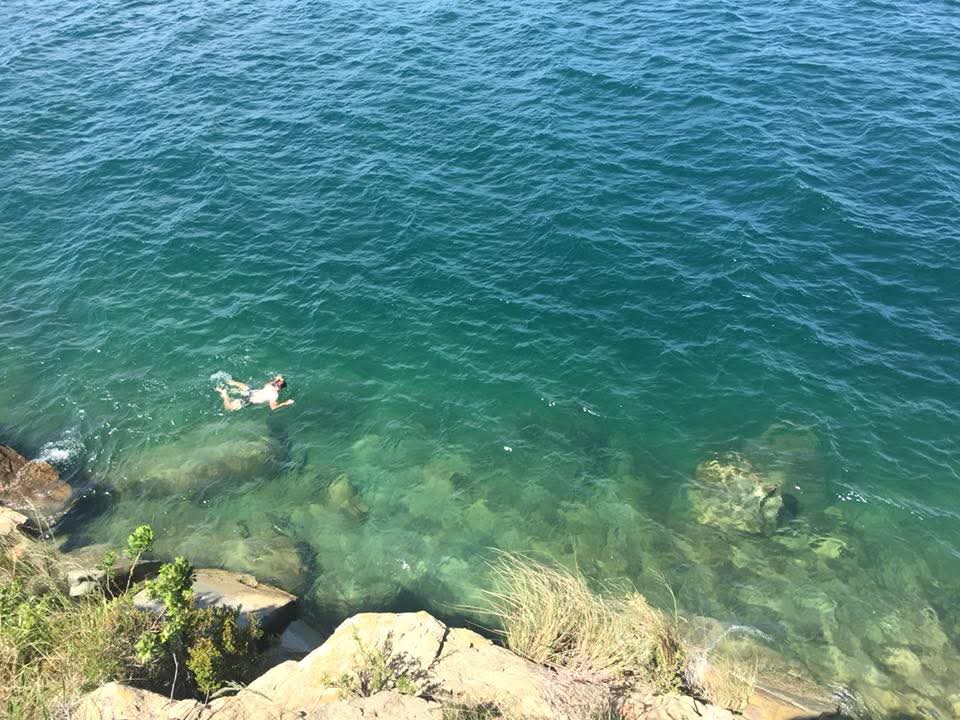 Angelo swimming out in clear turquoise water, seen from the rocky ledge above
