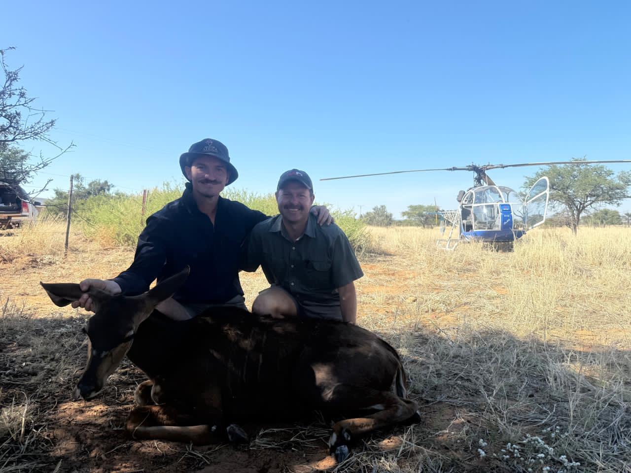 Angelo and Alwyn kneeling together beside an antelope with the helicopter behind them