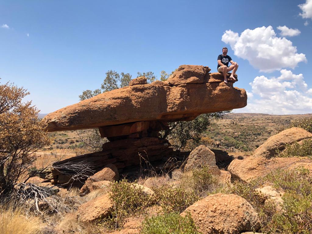 Angelo sitting on the edge of a balancing mushroom-shaped rock under a blue sky